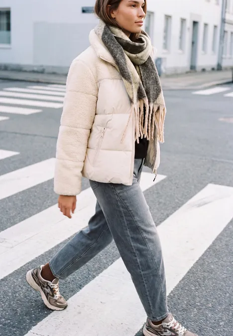 Frau überquert Straße im Winteroutfit: helle Daunenjacke, Schal, graue Jeans und braune Sneaker.