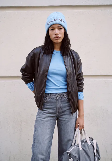 Frau in Lederjacke, blauer Mütze, Jeans und Tasche steht vor heller Wand.