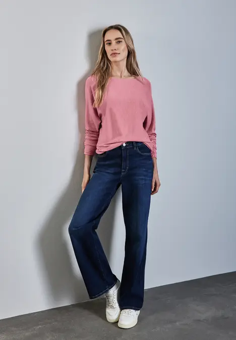 Frau mit rosa Pullover, dunkelblauer Jeans und weißen Sneakers steht an einer Wand.