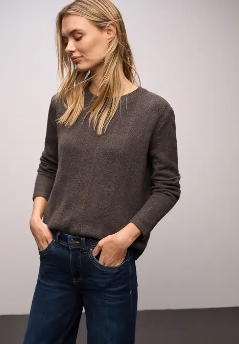 Frau in schlichtem grauen Pullover und blauen Jeans steht lässig vor neutralem Hintergrund.