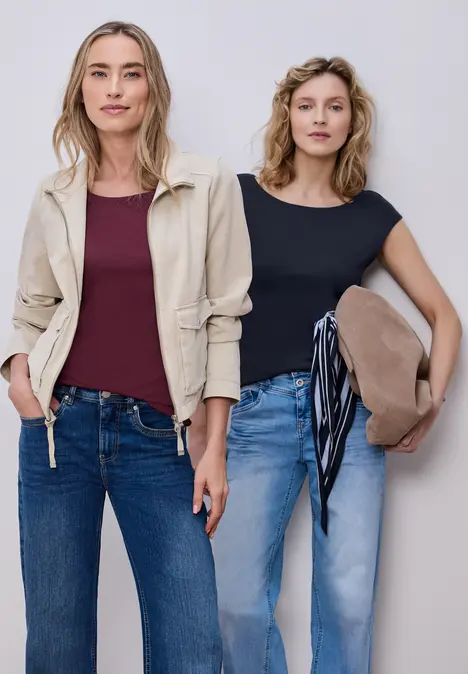 Zwei Frauen in Jeans stehen nebeneinander, eine mit beiger Jacke und rotem Shirt, die andere im schwarzen Top mit Hut.
