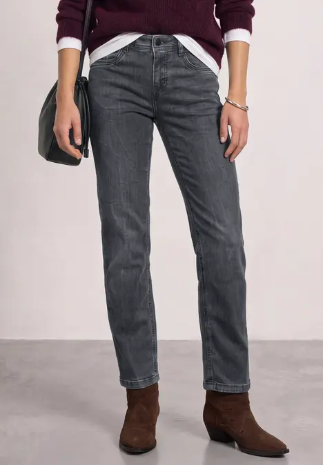 Eine Person trägt dunkelgraue Jeans und braune Ankle-Boots zu weißem Shirt und bordeauxrotem Cardigan.