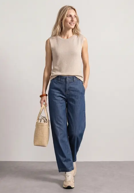 Eine Frau in beigem Top, weitem blauen Jeans und Sneakers, hält eine Basttasche vor hellem Hintergrund.