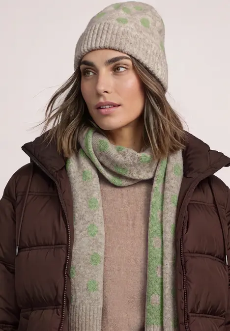 Frau mit grauer Mütze und grün gepunktetem Schal, braune Daunenjacke.