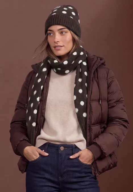 Frau trägt eine braune Steppjacke, beigen Pullover, blaue Jeans, gepunkteten Schal und Mütze