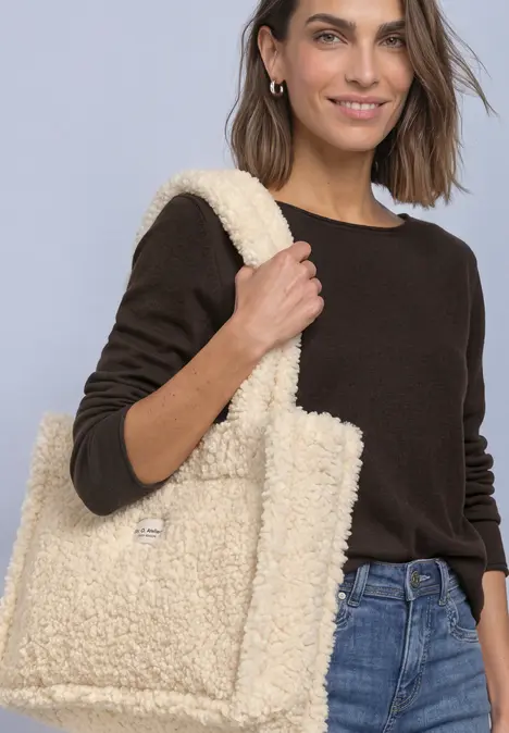 Eine Frau in schwarzem Pullover und Jeans trägt vor hellem Hintergrund eine große, flauschige beige Teddy-Tasche.