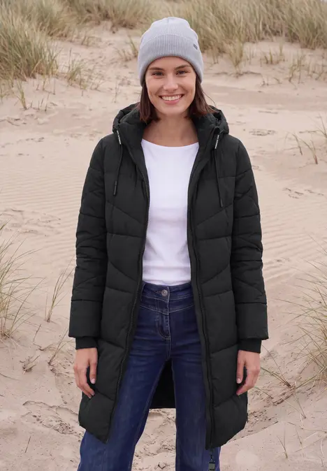 Frau in schwarzer Steppjacke mit grauer Mütze steht am Strand vor Dünen.
