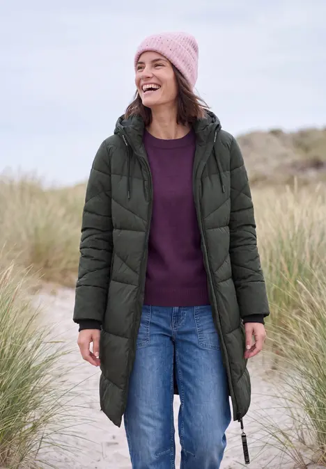 Eine lächelnde Frau in grüner Steppjacke mit Kapuze, rosa Mütze, Pullover und Jeans im Dünengras.