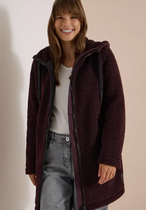 Eine lächelnde Frau trägt einen dunklen, langen Teddy-Fleece-Mantel mit Kapuze und Reißverschluss.