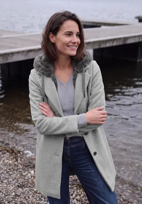 Frau in grauem Mantel und Jeans steht am Ufer, lächelt verschmitzt mit verschränkten Armen vor einem Steg.