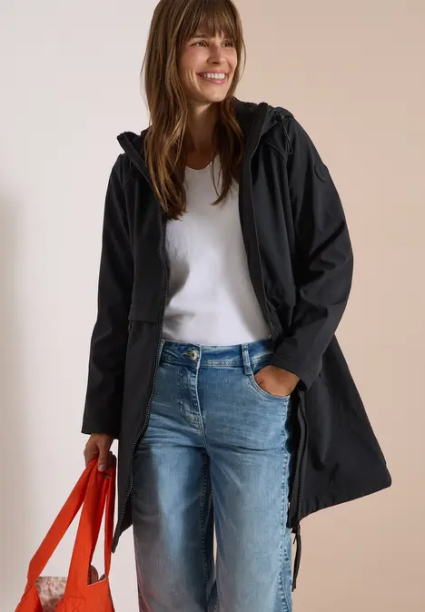 Une femme sourit en manteau à capuche noir, pull blanc et jeans, tenant un sac rouge.