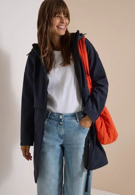 Femme en jean, t-shirt blanc et manteau bleu foncé, portant un sac orange de manière décontractée.