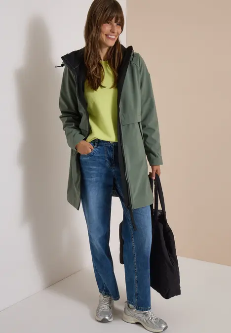 Une femme souriante en manteau vert, jean et baskets, tenant un sac noir.