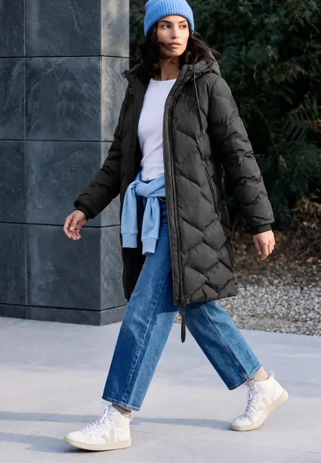 Frau spaziert in langer, gesteppter Daunenjacke mit Kapuze, Jeans, Sneakern und Mütze.