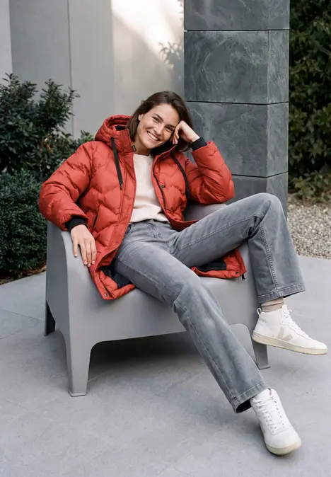 Frau in roter Winterjacke und grauer Jeans sitzt entspannt auf Stuhl und lächelt.