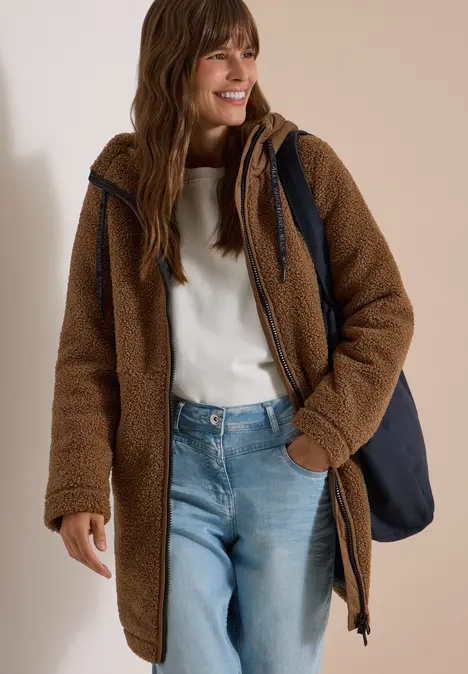 Eine lächelnde Frau trägt eine braune Teddyjacke, weißen Pullover, blaue Jeans und eine dunkelblaue Tasche.