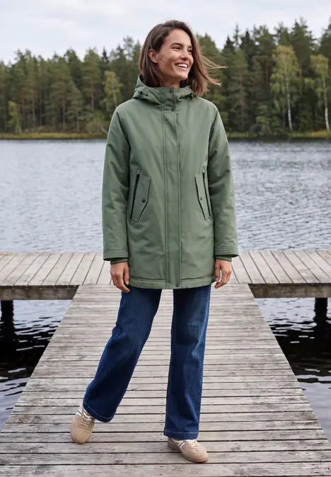 Frau in grünem Parka steht lächelnd auf einem Holzsteg am See, umgeben von Bäumen.