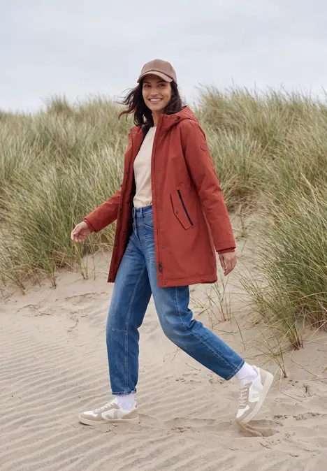 Frau in roter Jacke, Jeans und Cap spaziert lächelnd mit weißen Schuhen über einen sandigen Dünenstrand mit Gras.