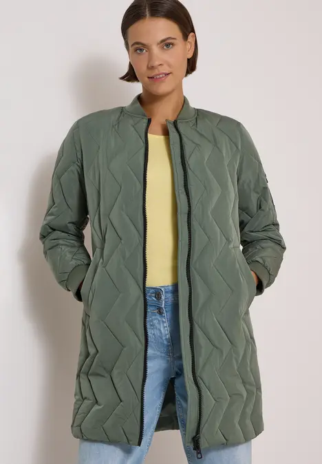 Eine Frau in einem langen, olivgrünen, gesteppten Mantel mit Zickzack-Design, gelbem Top und Jeans.