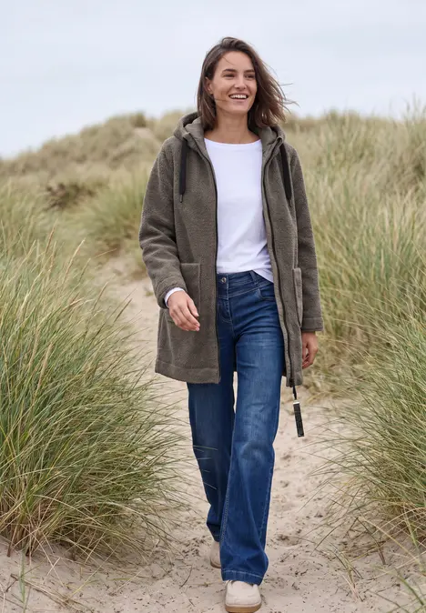 Femme souriante en manteau vert et jean marchant dans les dunes au bord de la mer.