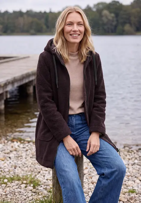 Eine Frau in brauner Jacke sitzt entspannt auf Steinen am Ufer, im Hintergrund ein Steg und ruhiger See.