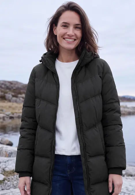 Eine lächelnde Frau in schwarzer Steppjacke steht an einem felsigen Strand.