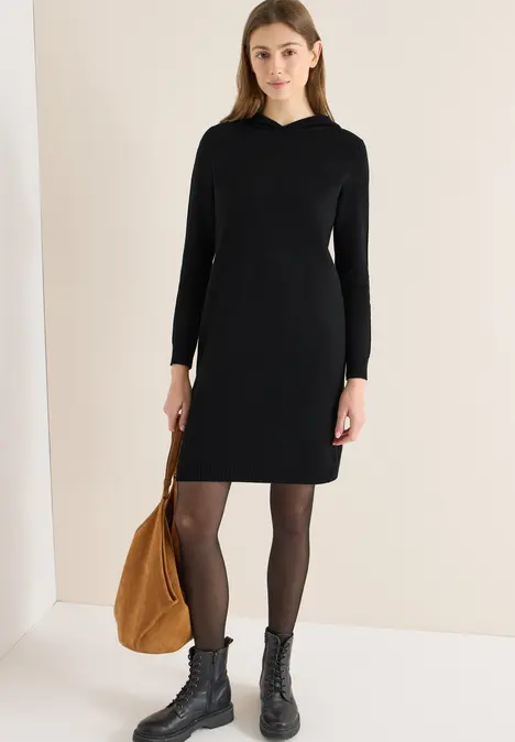 Vrouw in zwarte jurk met lange mouwen, zwarte panty's en laarzen, met een bruine tas.