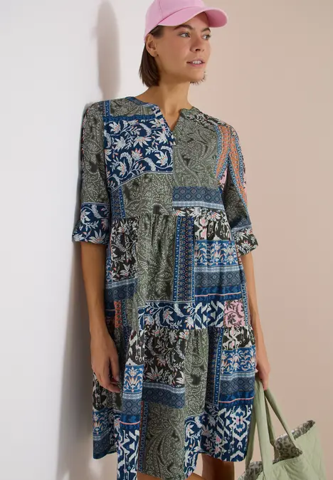Frau in blau-grünem Patchwork-Kleid mit 3/4-Ärmeln, rosa Kappe und Netz-Einkaufstasche.