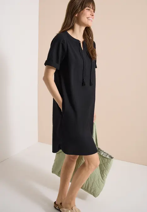 Eine Frau in einem lässigen schwarzen Kurzarmkleid mit Taschen und Quasten vor hellem Hintergrund.