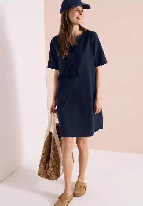Frau in marineblauem T-Shirt-Kleid mit Cap, Korbtasche und Slippern vor hellem Hintergrund.