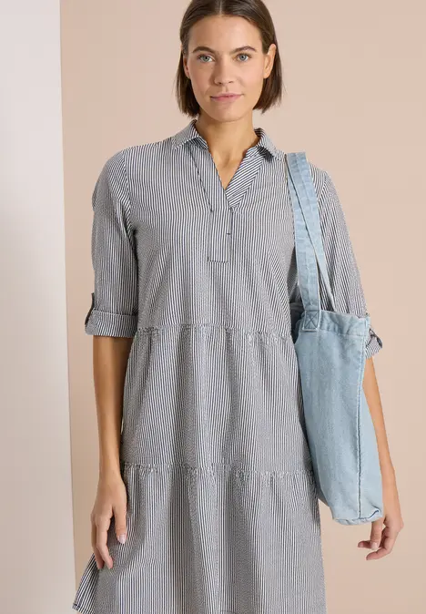 Frau in hellgrauem, knielangem Hemdkleid mit 3/4-Ärmeln und Jeans-Schultertasche.