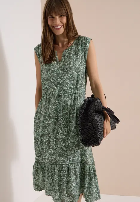 Femme porte une robe longue verte sans manches à motif floral, ceinturée, tenant un sac rond en paille.