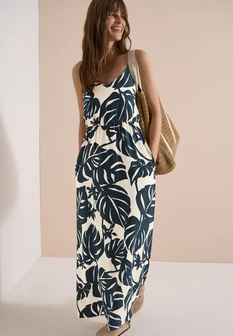 Femme en robe longue estivale à motifs de feuilles bleues, porte un panier en osier.