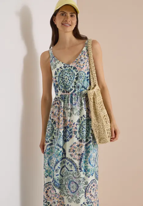 Eine lächelnde Frau trägt ein bunt gemustertes, ärmelloses Maxikleid, eine Strohtasche und eine gelbe Mütze.