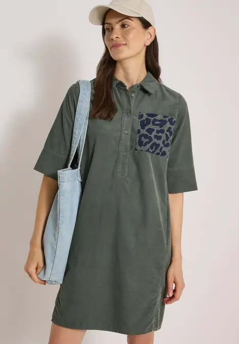 Vrouw in olijfgroene polo jurk met luipaardzakje, beige pet en lichtblauwe canvas tas over de schouder.