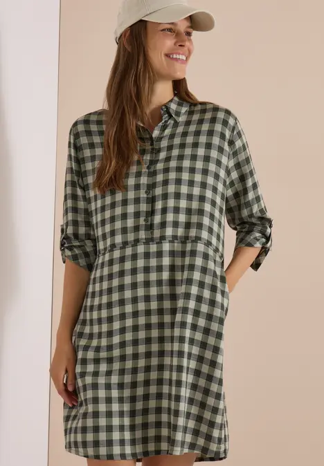 Frau in grün-schwarz kariertem Hemdkleid mit aufgekrempelten Ärmeln und Taschen, lächelnd mit Baseballcap.