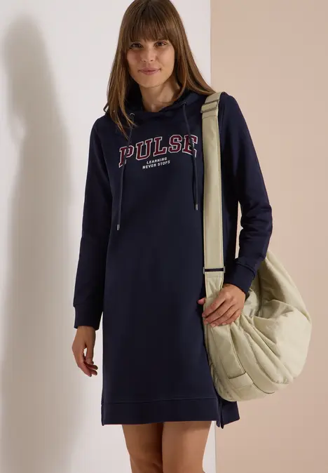 Frau im dunkelblauen Hoodie-Kleid mit "PULSAR"-Schriftzug, hält eine große, gesteppt beigetönte Umhängetasche.