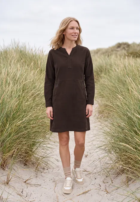 Frau in einem langen braunen Kleid steht an einem sandigen Strand mit Dünen