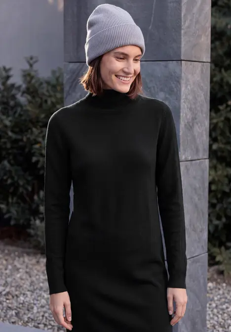 Eine lächelnde Frau in schwarzem Rollkragenkleid und grauer Mütze steht draußen neben einem Steinpfeiler und Pflanzen.