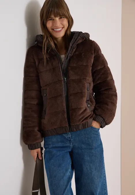 Junge Frau lacht, trägt braune Steppjacke, blaue Jeans und hält eine Tasche.