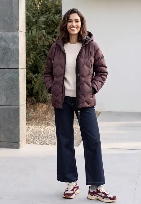 Eine lächelnde Frau in gepolsterter Jacke, hellem Pullover, weitem Jeans und Sneakern im Freien.