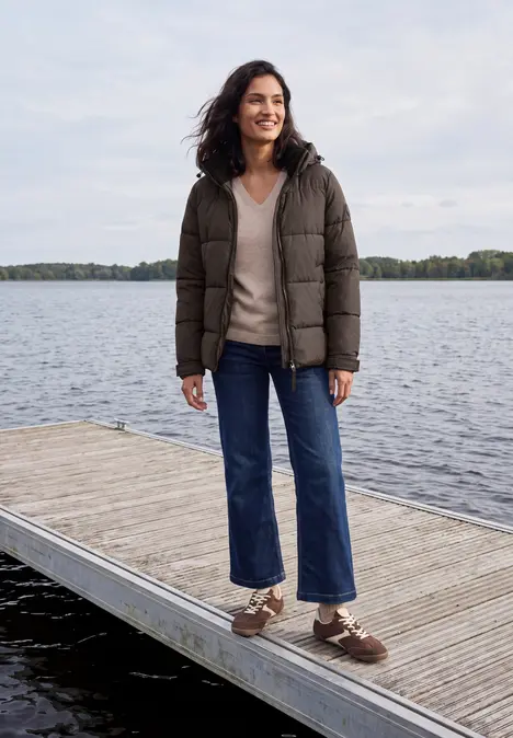 Eine Frau in dunkler Daunenjacke und weiten Jeans steht auf einem Steg am See und lächelt.