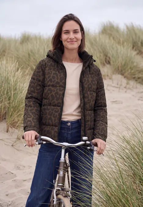 Eine lächelnde Frau mit Fahrrad in einer Dünenlandschaft, trägt Daunenjacke, Jeans und T-Shirt.