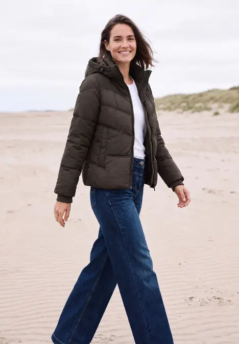 Frau in Winterjacke und Jeans spaziert am Strand, lächelt in die Kamera