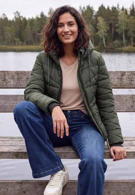 Eine lächelnde Frau in grüner Steppjacke sitzt auf einer Bank am See, im Hintergrund Wald.