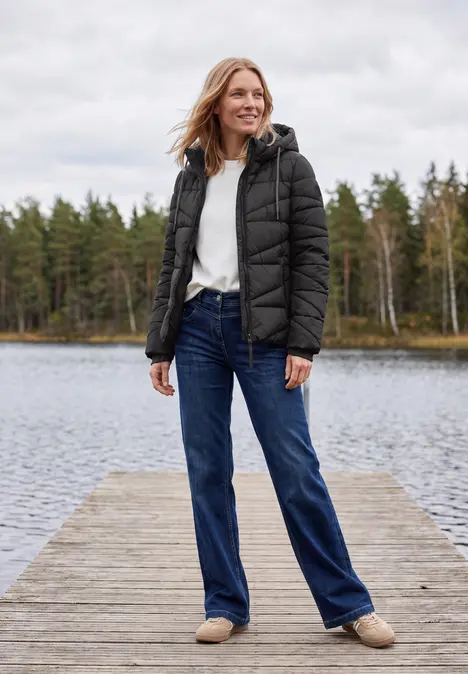 Frau in schwarzer Daunenjacke, weißem Pullover, blauen Jeans und Hausschuhen steht lächelnd auf einem Steg am See.
