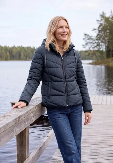 Une femme souriante en doudoune noire et jean sur une passerelle au bord d'un lac en forêt.