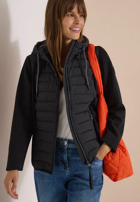 Frau in schwarzer Steppjacke, Jeans und weißem Top mit oranger Tragetasche vor hellem Hintergrund.