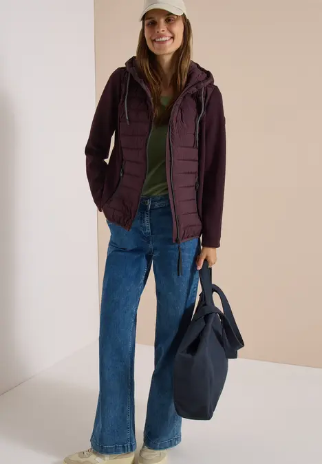 Eine Frau posiert in einer gesteppte Jacke, grünem Shirt, Schlagjeans, Cap und großer Tasche.