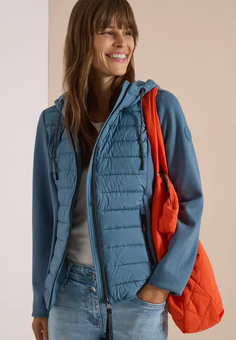 Lächelnde Frau in hellblauer Steppjacke mit Softshell-Ärmeln, Jeans und orangefarbener Tasche.